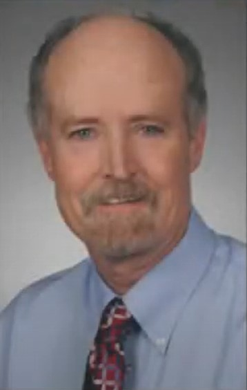 A person posing in front of a grey background. He is wearing a blue button down. 